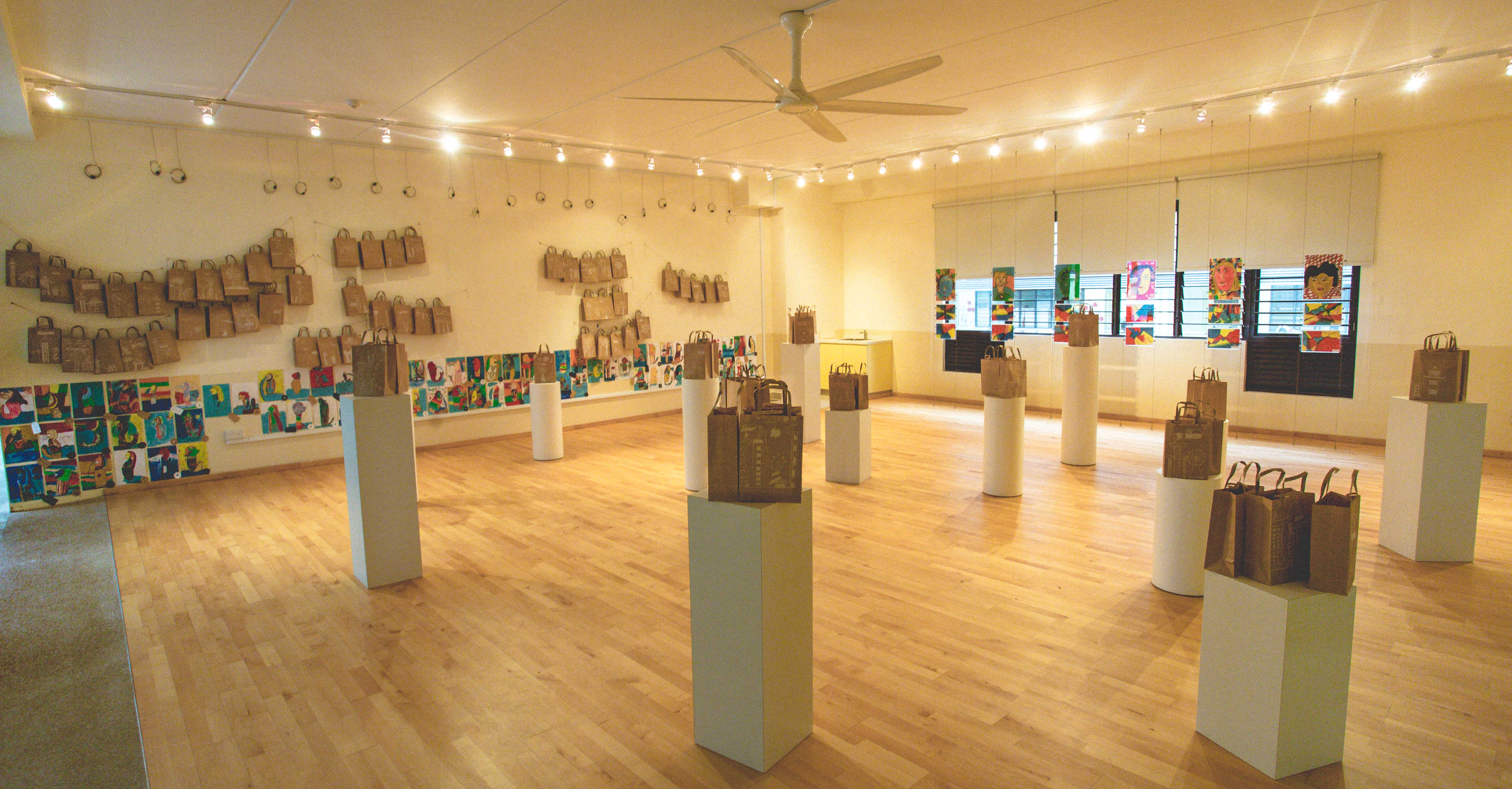 Gallery room with brown paper bags on walls and pedestals, colorful artwork displayed along the walls, wood floor.